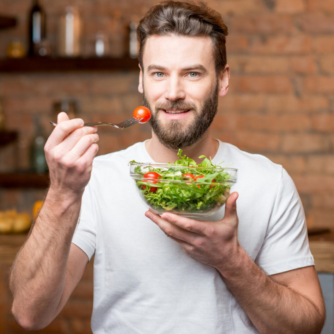 Handsome bearded man in white t-shirt eating salad with tomatoes in the kitchen. Healthy and vegan food concept